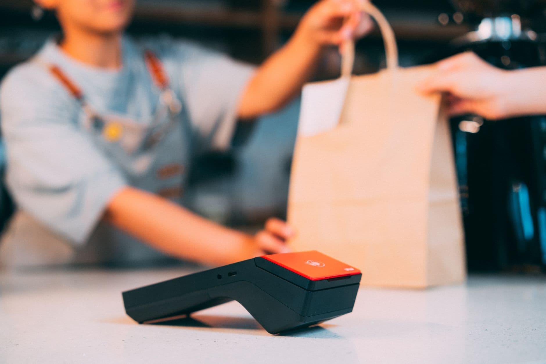 Cashier handing a takeaway bag to a customer with a contactless payment terminal in focus.