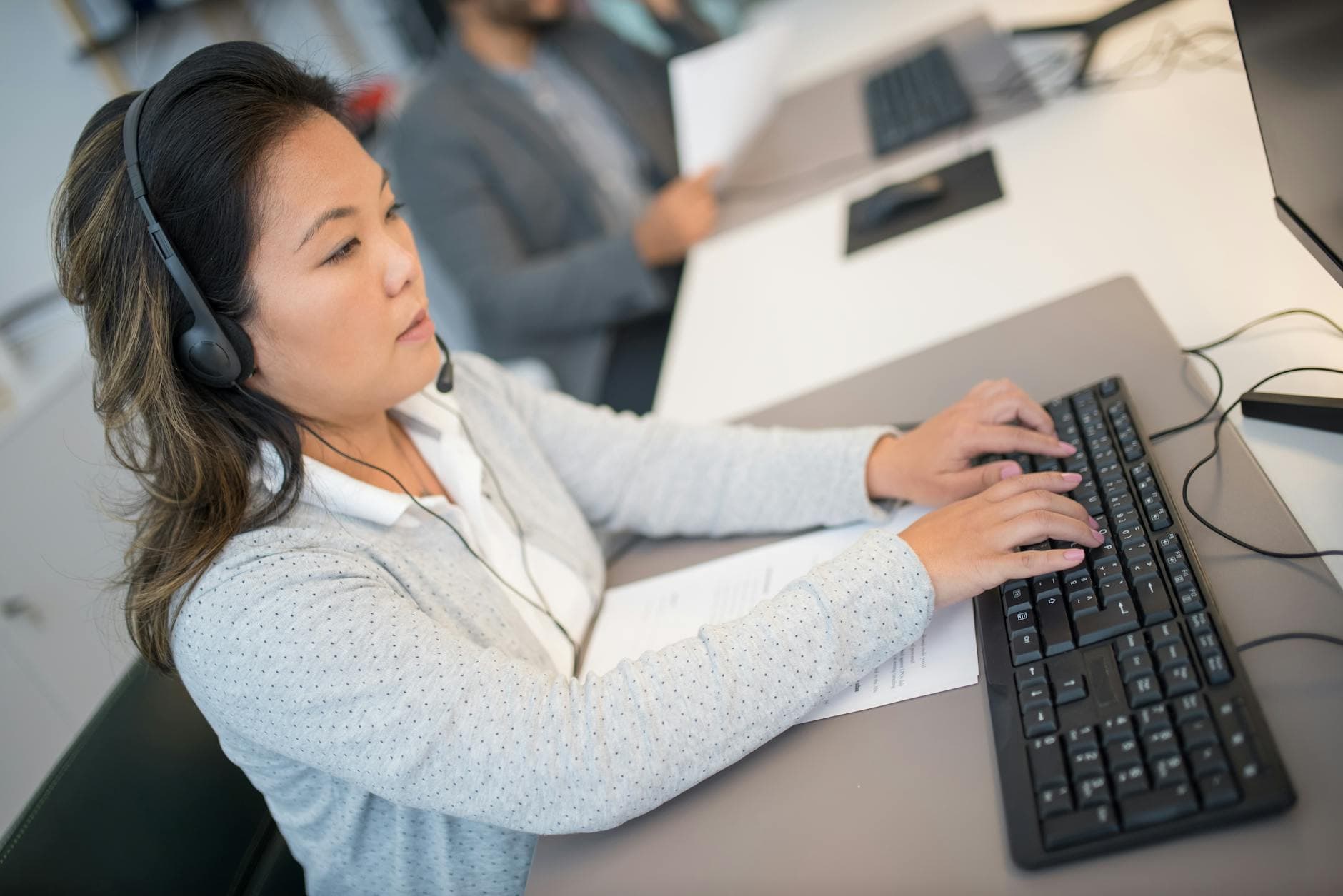 Asian woman working at a computer with headphones in a modern office setting.