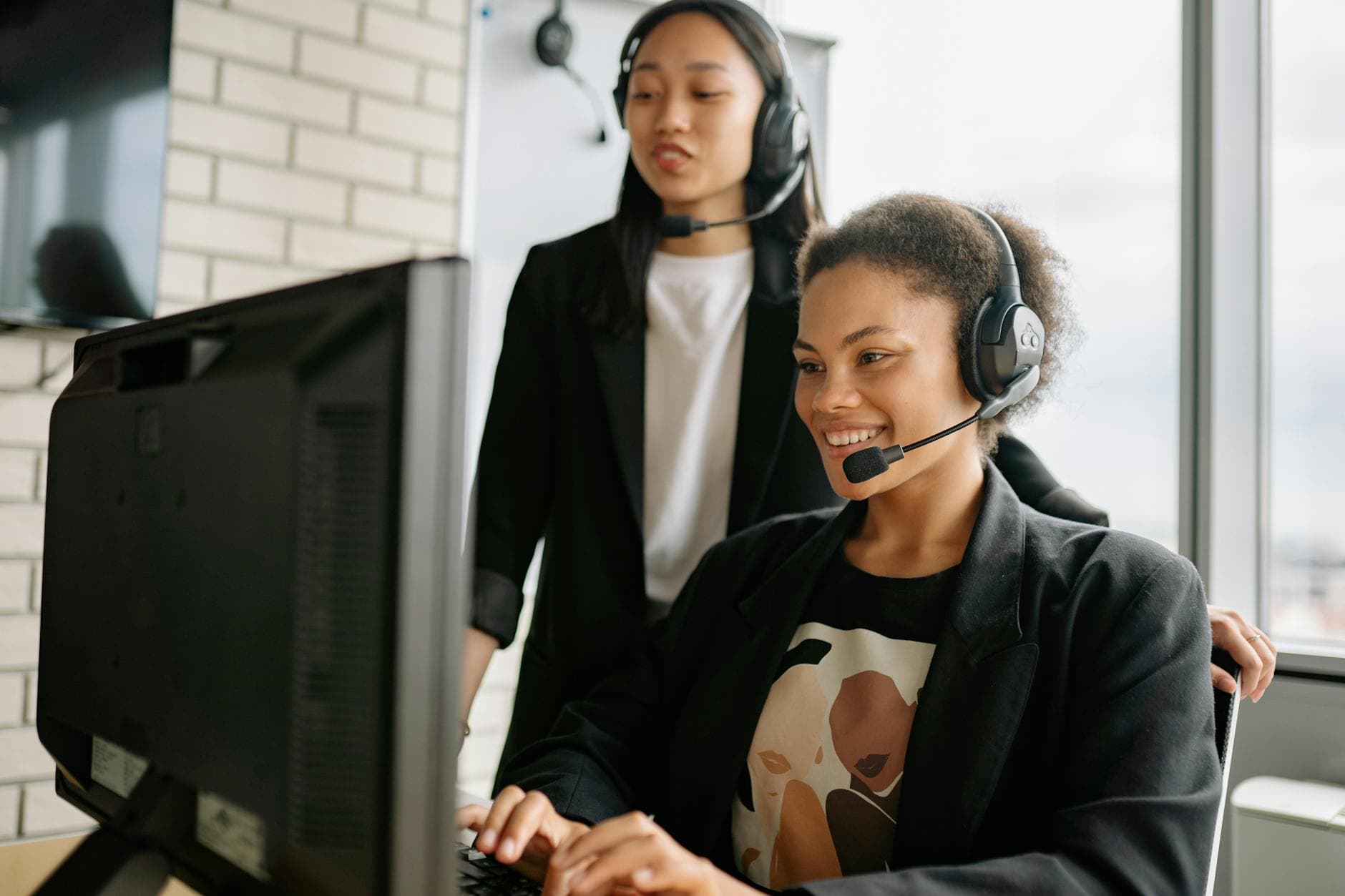 Two women with headsets engaged in customer service at a computer in a bright office.